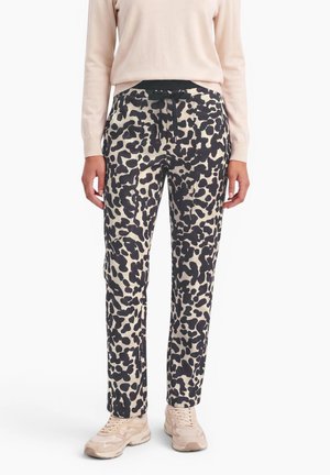 Mujer con pantalones negros y beige con manchas, camiseta beige de manga larga y zapatillas deportivas beige, de pie con las manos relajadas.