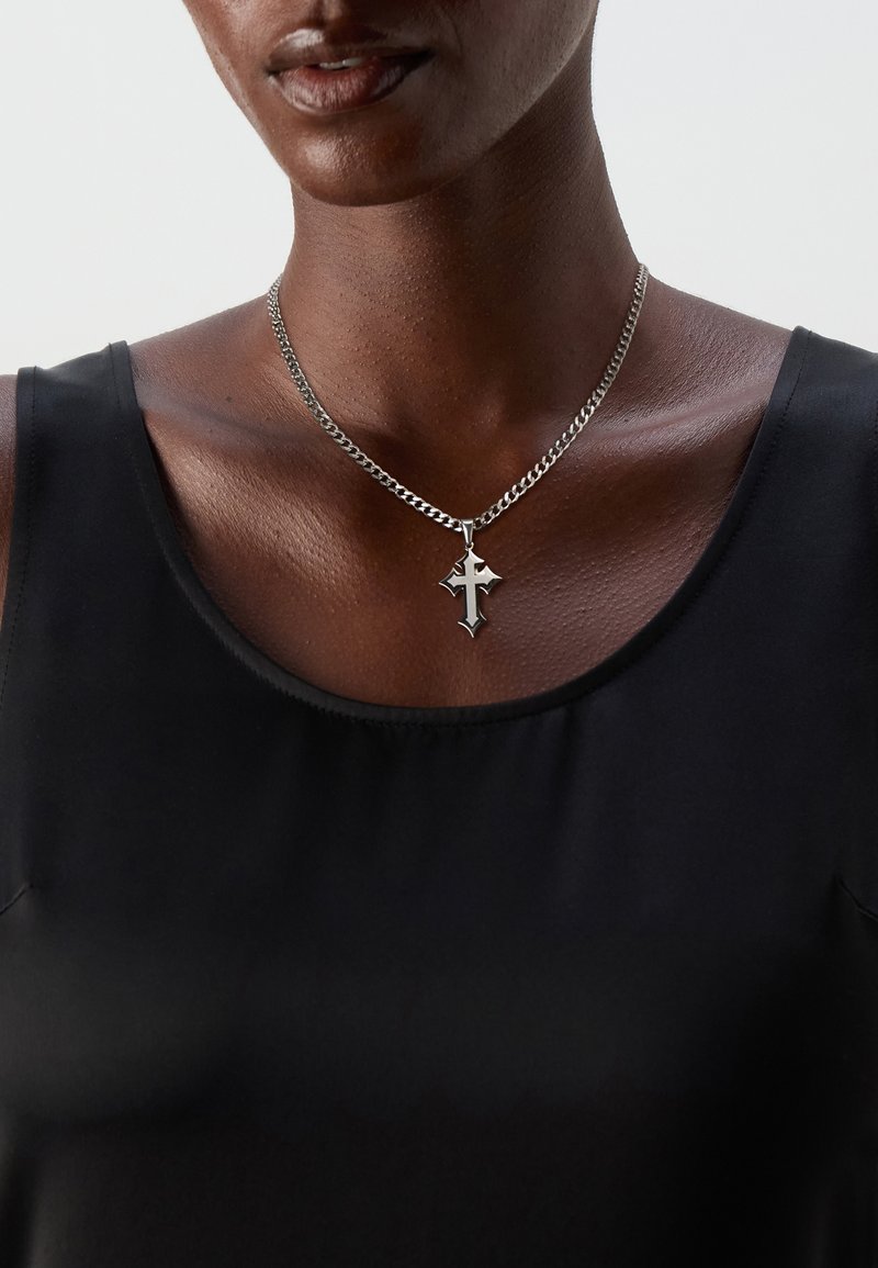 Silver chain necklace featuring a cross pendant with pointed edges, worn over a black sleeveless top, highlighting the shiny metal texture.