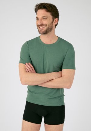T-shirt vert à manches courtes en tissu doux, col rond, porté avec un short de boxeur noir. Design simple sans motifs ni logos visibles.