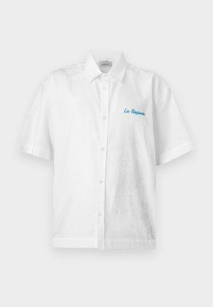 Joven de pie con una mano en el bolsillo, vestido con una camisa blanca de manga corta abotonada, pantalones cortos negros, calcetines blancos y zapatillas negras altas.