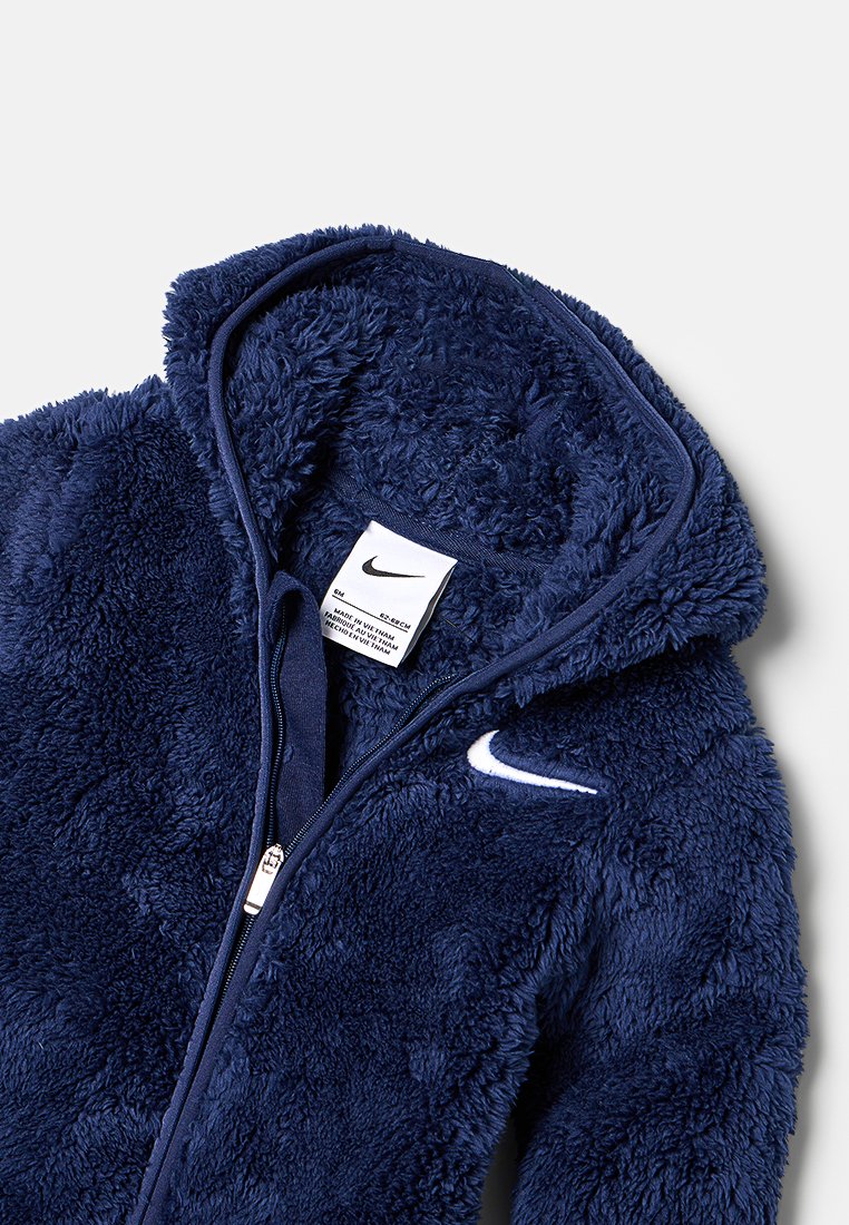 Marinblå fleecejacka med huva som har en mjuk textur, full dragkedja och en vit Nike-logotyp på bröstet. Fodrad insida med skötselråd synligt.