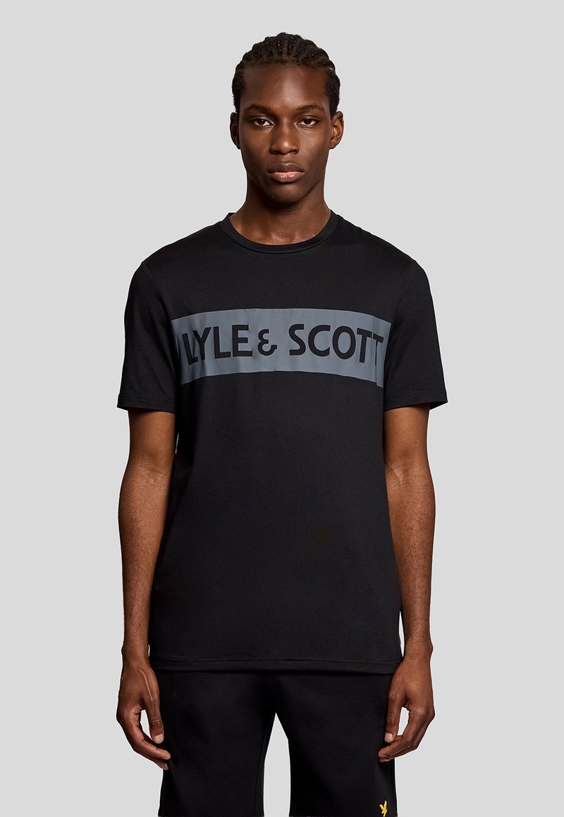 Jeune homme aux cheveux tressés portant un t-shirt noir Lyle & Scott et un short noir, debout devant un fond gris uni.