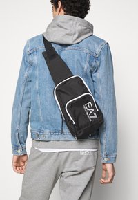 Borsa a tracolla nera realizzata in tessuto resistente. Caratterizzata da un compartimento principale con zip, dettagli bianchi e logo "EA" sul davanti.