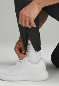 Sorte atletiske bukser med lynlås ved anklerne, båret med hvide sneakers. Stoffet ser let ud med en glat tekstur.