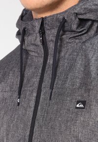 Grå herringbone-jacka med hög krage, svarta dragsnören och diskret logotyp-badge. Har en full framdragkedja och texturerad tyg.