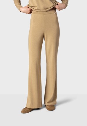 Une personne ajuste un pantalon beige à jambes larges assorti à des chaussures coordonnées, sur un fond gris uni.