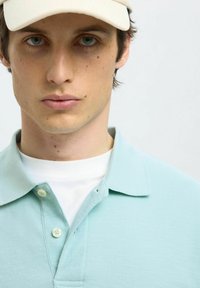 Ljust blå pikétröja med strukturerad yta, tvåknappsslutning och spetsig krage, över en vit T-shirt. Kepa i neutral färg.