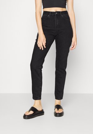 Femme portant un jean noir taille haute, un crop top noir et des sandales à plateforme noires, debout devant un fond clair uni.