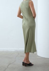 Robe midi vert olive avec une silhouette ajustée, design sans manches et fermeture éclair au dos. Portée avec des sandales à talons blocs noires.