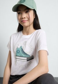 T-shirt blanc avec un graphisme d'une sneaker Converse teal. Le modèle porte une casquette vert clair et un pantalon foncé. Tissu doux, design décontracté.