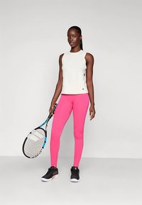 Fehér ujjatlan felső pink bordázott leggingsszel és fekete tornacipővel; kék díszítésű teniszütőt tart a kezében.