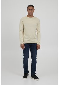 Beige langermet genser laget av strikket stoff, med en teksturert frontpanel og rund halsutringning. Kombinert med mørkeblå jeans og svarte joggesko.