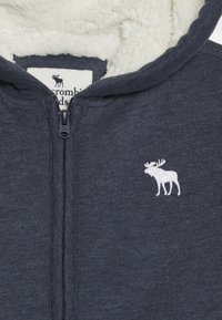 Felpa grigia con zip, foderata in pile e logo ricamato di un alce bianco sul lato sinistro del petto. Soft texture e design aderente per il calore.