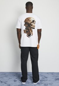 White t-shirt featuring a 3D wooden skull design on the back. Paired with black sweatpants and a tag on the side. Smooth fabric texture.