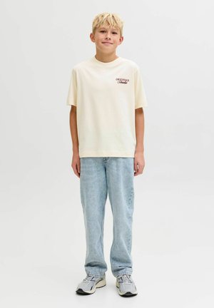 Crème katoenen t-shirt met de tekst "ORIGINALS" in bordeauxrood op de linkerborst, gecombineerd met lichte baggy jeans en grijze sneakers.