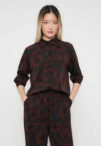 Jeune femme aux longs cheveux lisses, portant une chemise à boutons sombre et un pantalon à grand motif floral, les mains dans les poches, fond neutre.