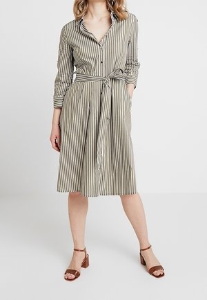 Nainen, jolla on polvipituinen, pitkähihainen, beige-valkoraidallinen paitamekko, jossa on vyötärösolmu ja ruskeat paksukorkoiset sandaalit.