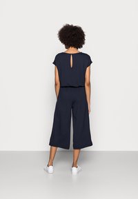 Granatowy kombinezon z krótkimi rękawami, detalem w kształcie dziurki na plecach oraz szerokimi nogawkami. Wykonany z miękkiego materiału, w zestawieniu z białymi trampkami.