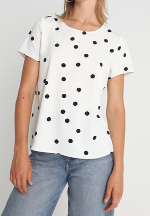 Kobieta ubrana w biały top z krótkim rękawem w czarne kropki oraz jasnoniebieskie dżinsy, stojąca na jednolitym tle.