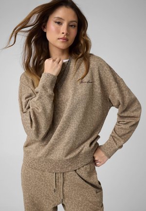 Ung kvinde med langt hår iført en beige sweatshirt med dyreprint og matchende bukser, poserer med den ene hånd nær hagen og den anden på hoften.