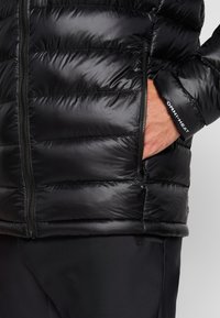 Sort puderjakke med en blank finish, der har lodrette kanaler, en lynlås lomme og "OMNI-HEAT" label på manchetten. Blød tekstur.