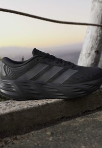 Black Adidas running shoe with three stripes and thick cushioned sole resting on a concrete ledge outdoors at sunset.