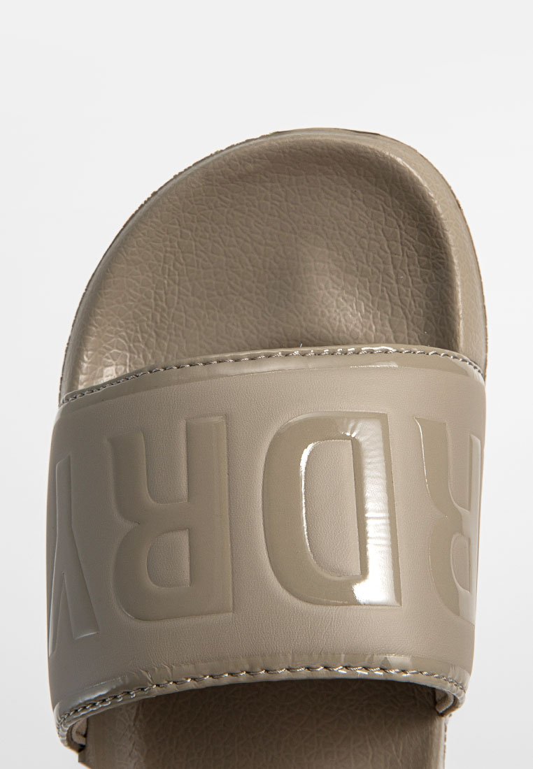 Sandale glissière beige avec une sangle texturée arborant un logo brillant. La semelle est plate, fabriquée en matériau synthétique avec une finition lisse.