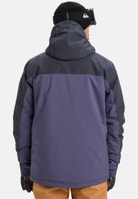 Man wearing a two-tone purple and dark gray winter jacket with a hood, black beanie, ski goggles, gloves, and brown pants facing away.