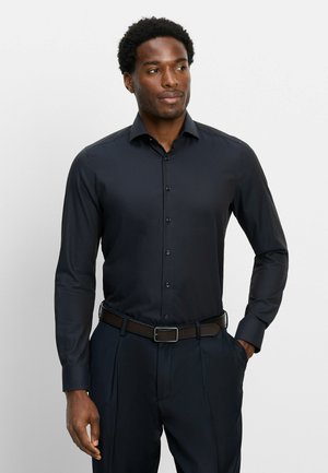 Man draagt een nauwsluitend zwart overhemd met knopen en zwarte broek, staat met één hand in de zak en kijkt opzij tegen een effen achtergrond.