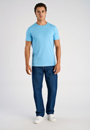 Mężczyzna stojący, ubrany w jasnoniebieską koszulkę z krótkim rękawem, ciemnoniebieskie dżinsy i białe trampki na jednolitym tle.