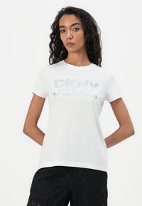 Weißes Baumwoll-T-Shirt mit einem metallisch silbernen "DKNY JEANS"-Logo, Rundhalsausschnitt und kurzen Ärmeln, kombiniert mit strukturierten schwarzen Shorts.