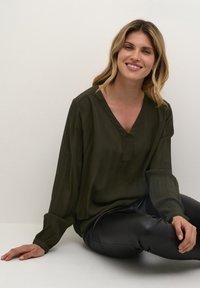 Mörkgrön långärmad blus med V-ringning och plisserad textur, i kombination med svarta figurnära byxor. Modell som sitter mot en ljus bakgrund.