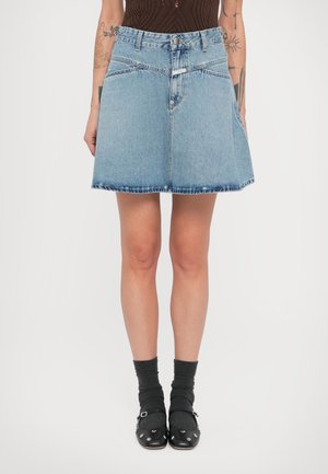 Jupe trapèze en denim bleu clair avec fermeture à boutons, poches et ourlet effiloché. Associée à des chaussures noires et des chaussettes grises.