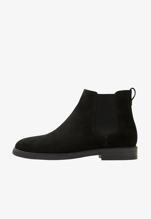 Bota Chelsea de ante negra con paneles laterales elásticos, un tirador en la parte trasera y una suela plana de goma con diseño de punta redonda.
