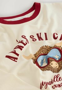 Cremefarvet sweatshirt med en burgunder krave og grafik af en brun bjerggækker i solbriller, med teksten "APRÈS SKI CHILL".