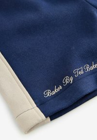 Tumedat sinine kangas koos beeži aksendiga servas. Omab tikitud teksti "Baker By Ted Baker." Sile tekstuur, lõbus disain.