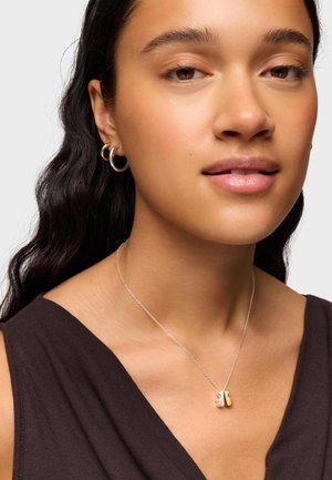 Donna con capelli lunghi e scuri che indossa orecchini a cerchio in oro e argento e una collana d'argento con un ciondolo a farfalla bicolore, vestita con una canotta scura.