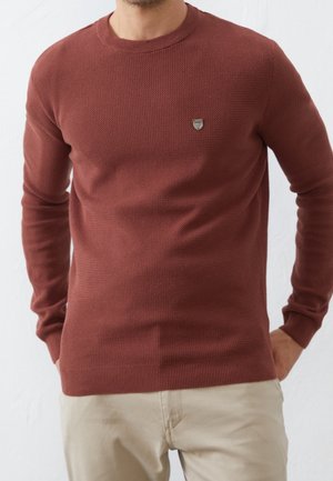 Homme portant un pull en maille texturée couleur rouille à manches longues et un pantalon beige, mains partiellement dans les poches, sur un fond uni.