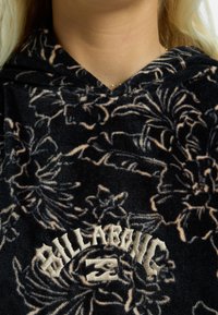 Sudadera con capucha negra con un patrón floral en beige. Presenta un logo bordado de "BILLABONG" en un diseño curvado en el pecho.