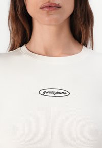 Rybkované, krémové priliehajúce tričko s okrúhlym výstrihom a vyšitým logom "guess.jeans" v čiernej farbe. Hladká textúra a tesný strih.
