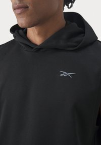 Fekete ujjatlan kapucnis pulóver, puha textúrával, elől zsebbel, zsinóros nyakkal és diszkrét szürke logóval a mellkason.