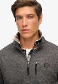 Donkergrijze fleecetrui met ritssluiting, van textuurstof, een geruite kraag in rood en blauw, en een klein logo op de borst.