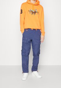 Orange hoodie with graphic design and drawstring, paired with navy cargo pants featuring multiple pockets and a belt, finished with white sneakers.