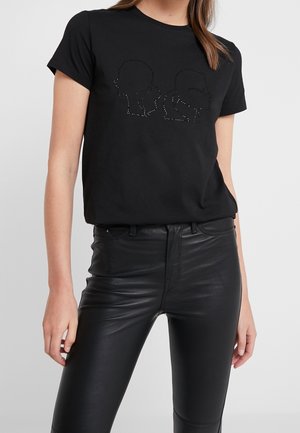 Kvinde iført en sort kortærmet skjorte med tre silhuetansigter i rhinsten, kombineret med højtaljede sorte læderbukser.