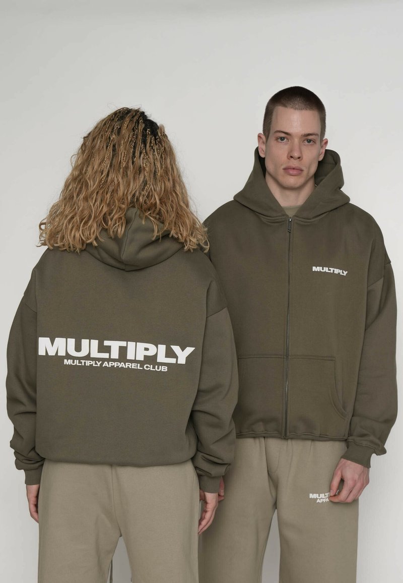 Sweat à capuche zippé vert olive avec un grand logo blanc "MULTIPLY" à l'avant et au dos. Le tissu semble doux ; porté avec un pantalon de survêtement beige.