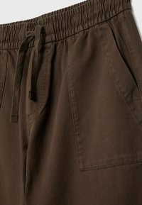 Gros plan sur un pantalon marron avec une ceinture élastique, des cordons de serrage et des poches latérales inclinées, fabriqué en tissu robuste.