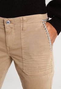 Pantalons en coton beige avec des clous en argent sur les côtés, poches avant et tissu texturé. La taille comprend une fermeture à bouton en argent.