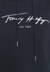 Felpa navy con logo bianco ricamato "Tommy Hilfiger" e testo "est. 1985". Presenta cordini con punte in metallo dorato. Texture del tessuto morbida.