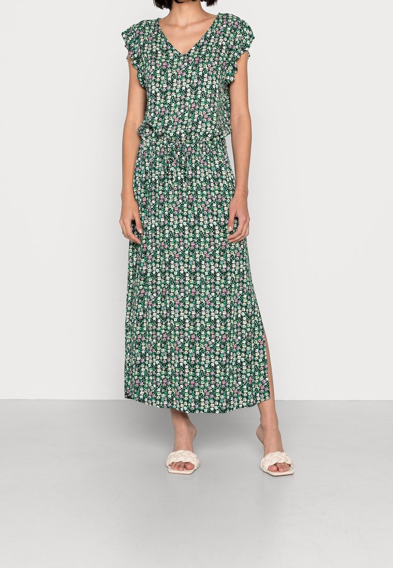 Femme portant une robe maxi verte fleurie avec des manches volantées et une fente latérale, associée à des sandales blanches à brides tressées.