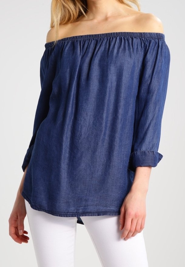 Femme portant un chemisier bleu marine à manches longues et épaules dénudées, associé à un pantalon blanc, sur un fond clair uni.